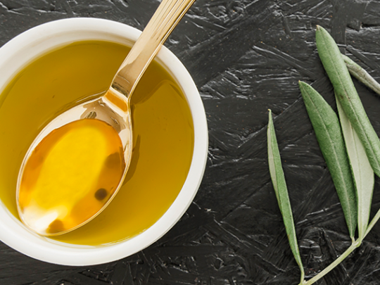 Bowl of olive oil with a spoon & an olive branch beside it.